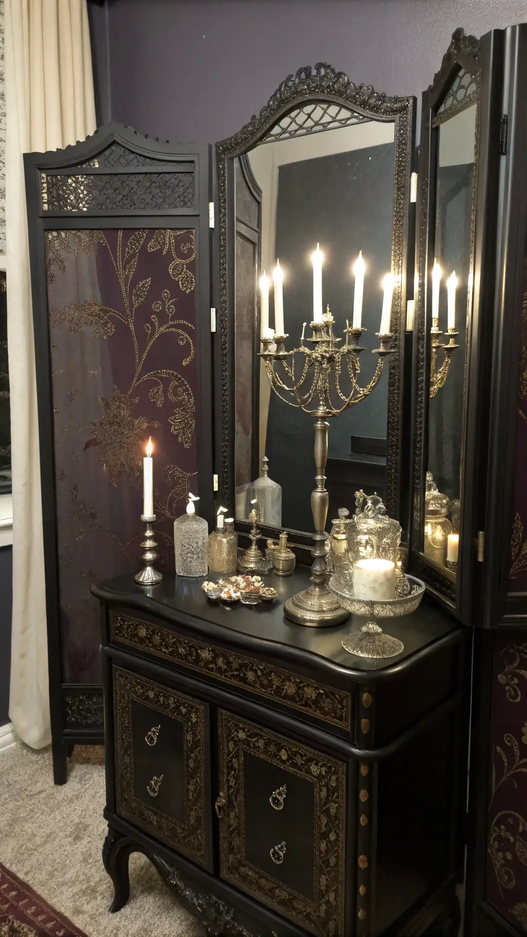 Dressing area with ornate black vanity, silver accessories, and aubergine brocade screen, lit by candlelight reflected in three-panel mirror