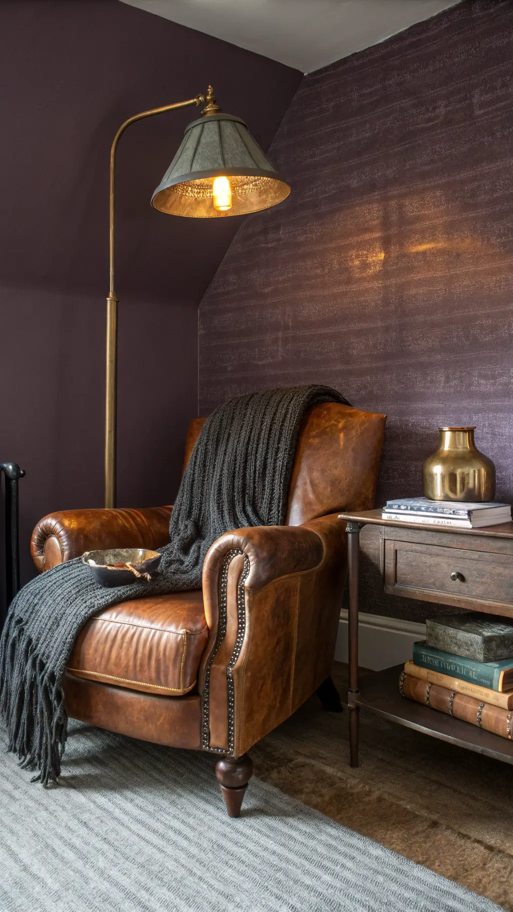 Low angle view of a cozy bedroom corner with a weathered leather armchair, chunky knit charcoal throw, antiqued brass floor lamp, and dark wooden side table with vintage books and a mercury glass vase against rich plum walls with textured grasscloth wallpaper, illuminated by golden hour natural light and warm LED fills.