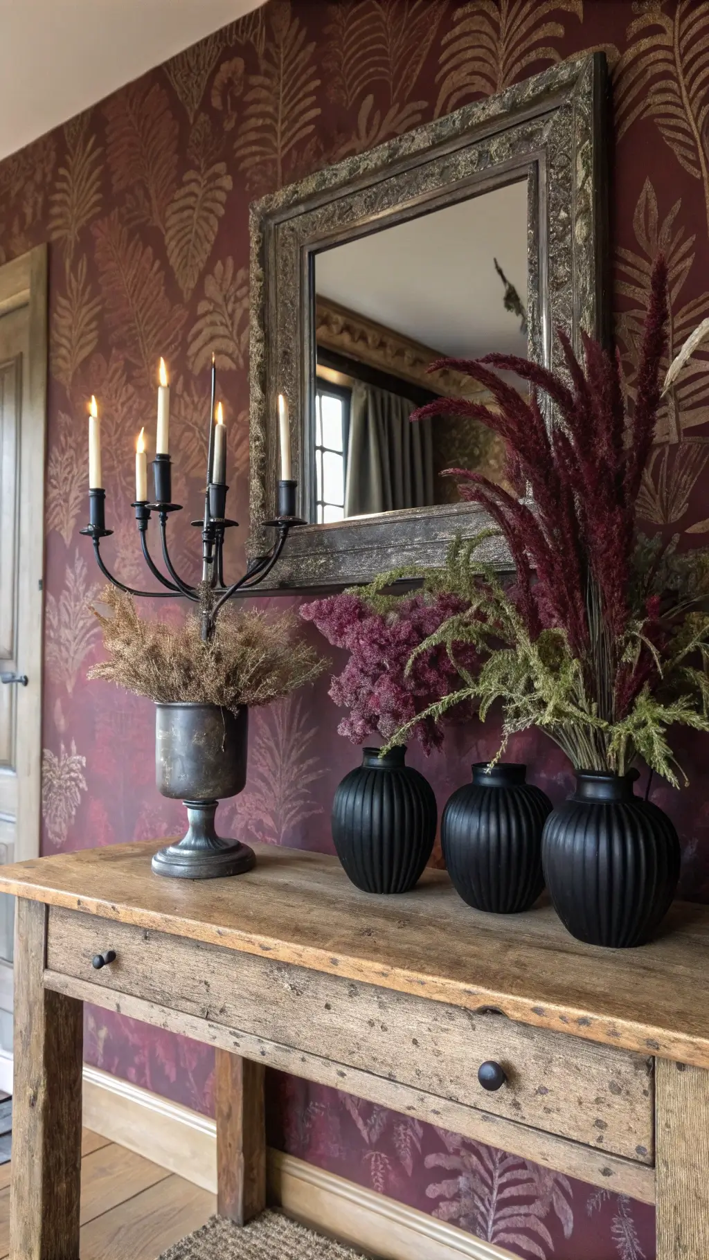 Antique oak console table against burgundy Morris & Co wallpaper with dried purple amaranthus and ferns in matte black vases, mirrored by a tarnished silver mirror reflecting candlelight for a mystical ambiance.