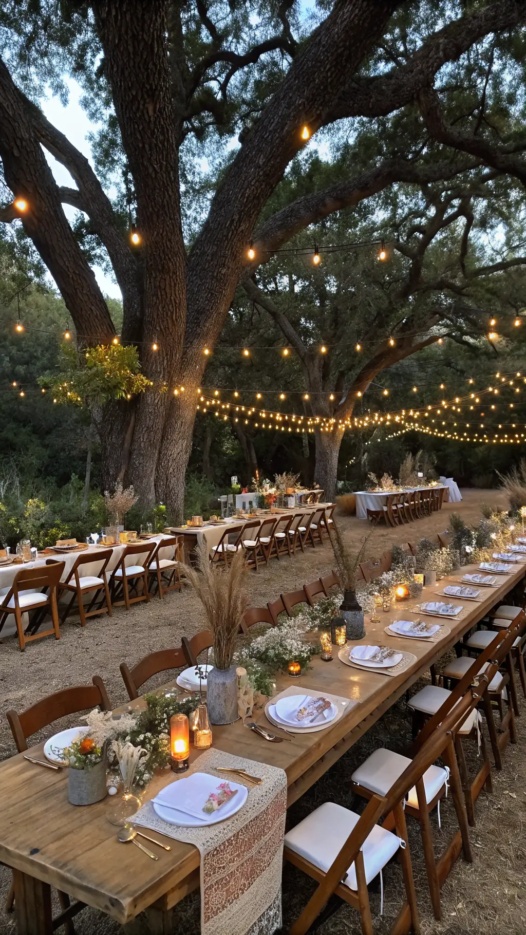 Outdoor dining setup under oak trees with string lights, long farm tables, wildflower centerpieces, mismatched vintage chairs, and terracotta napkins during blue hour.
