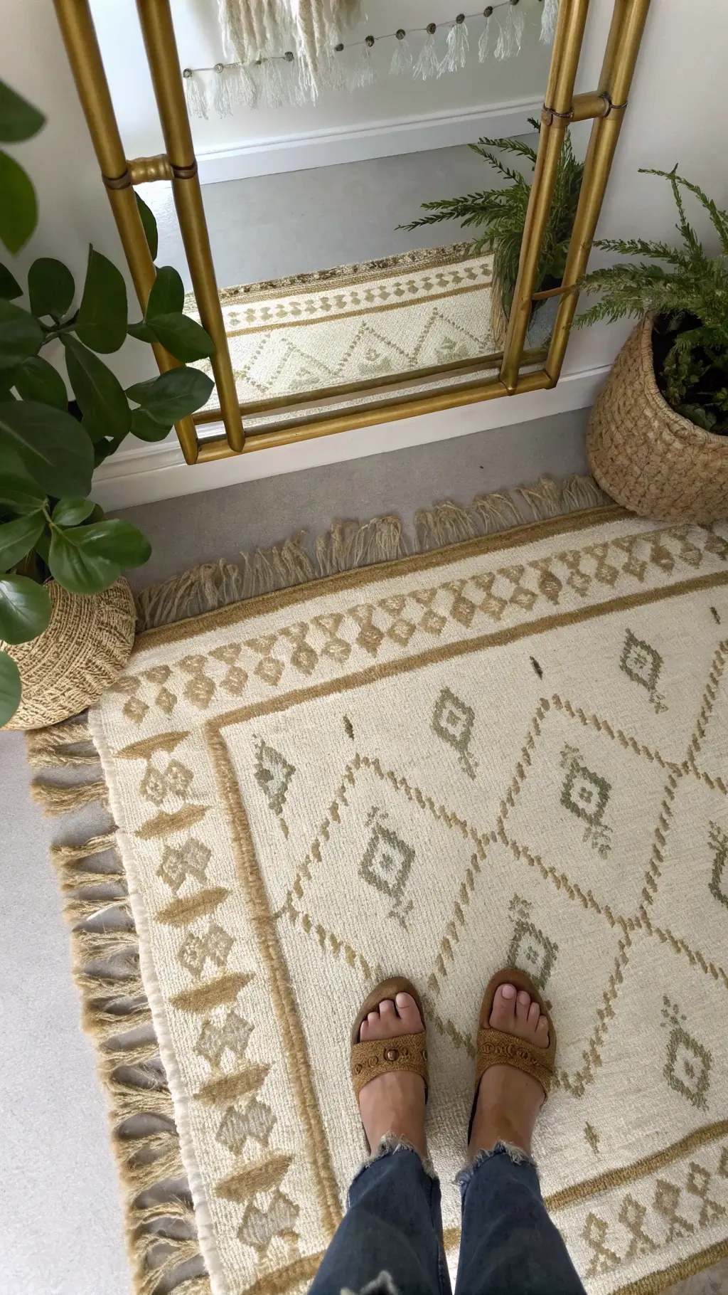 Layered jute and Moroccan wool rugs in cream and sage patterns, with feet of rattan bed frame and brass floor mirror visible. Green trailing plants frame the edges, giving off a grounded luxury mood.