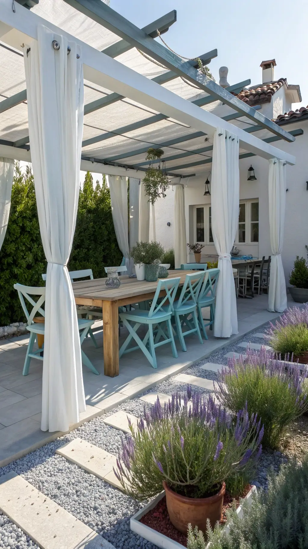 Midday view of a modern white-painted thrifted wooden dining set on a 10x15ft patio, surrounded by blue-gray gravel paths and clusters of potted lavender and rosemary, with mismatched pastel chairs under a pergola draped with white curtains.