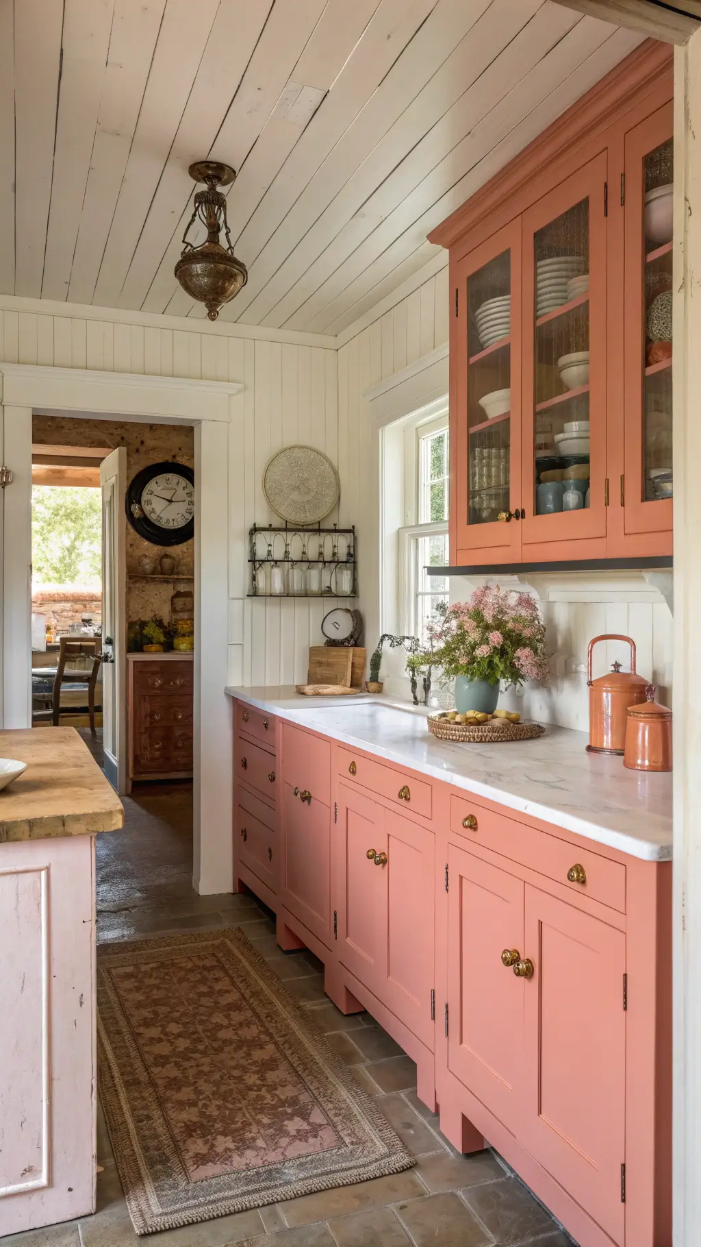 Cozy farmhouse-style kitchen with coral pink distressed cabinets, butcher block island, white counters, vintage copper handles, shiplap ceiling, antique kitchen scales, pottery, and dried flowers illuminated by late afternoon light.