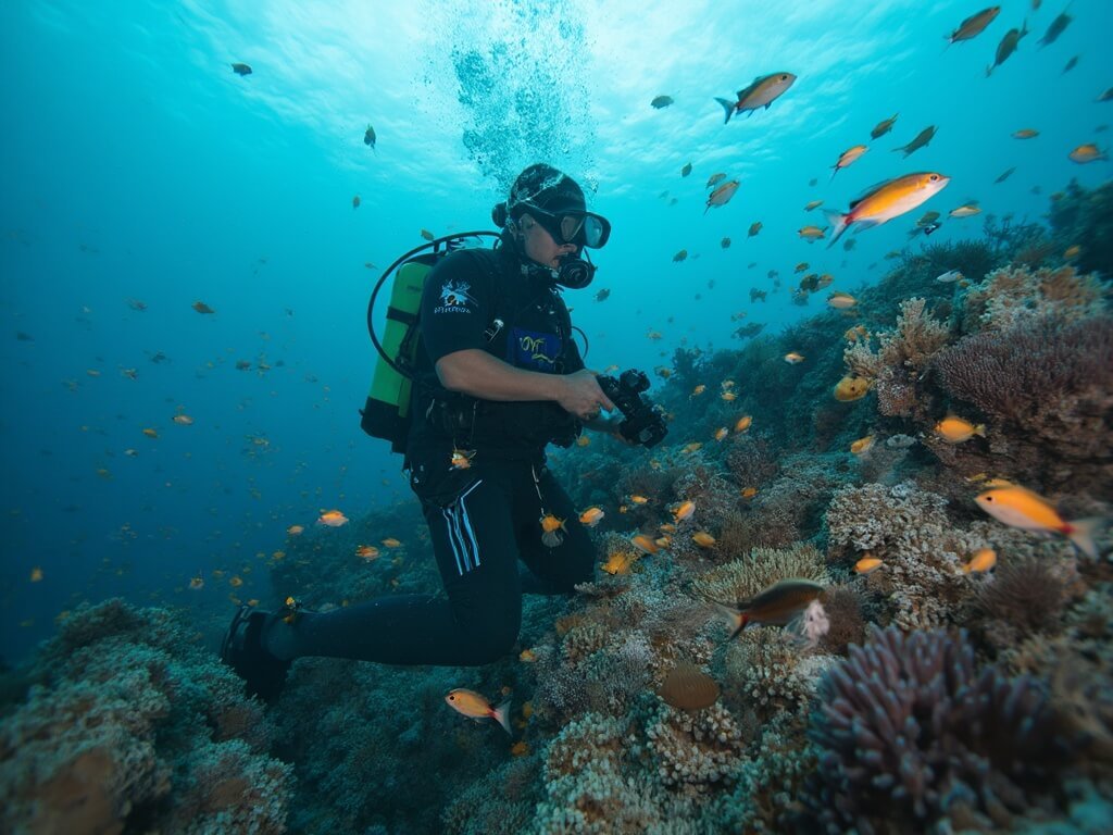 Papuan marine conservationist documenting marine biodiversity underwater amidst coral structures and colorful tropical fish while wearing professional diving gear