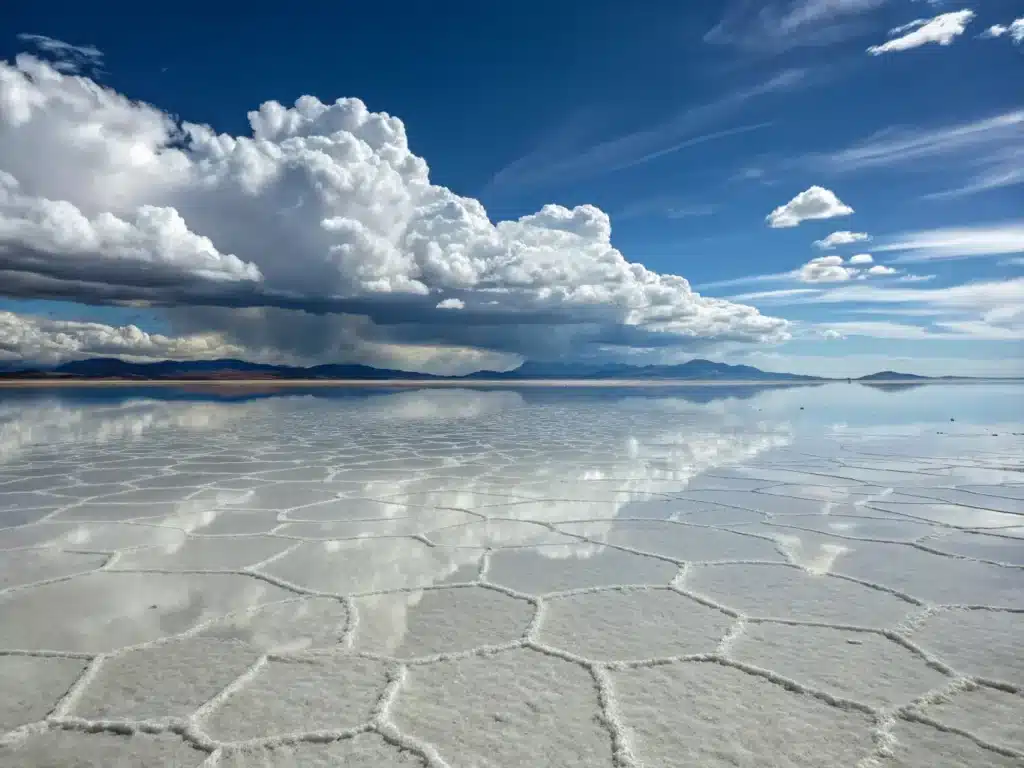 "Mirror-like reflection on Bolivia's Salar de Uyuni salt flat during rainy season with hexagonal salt patterns, distant Andean mountains, and sky duplicated in the crystalline water."