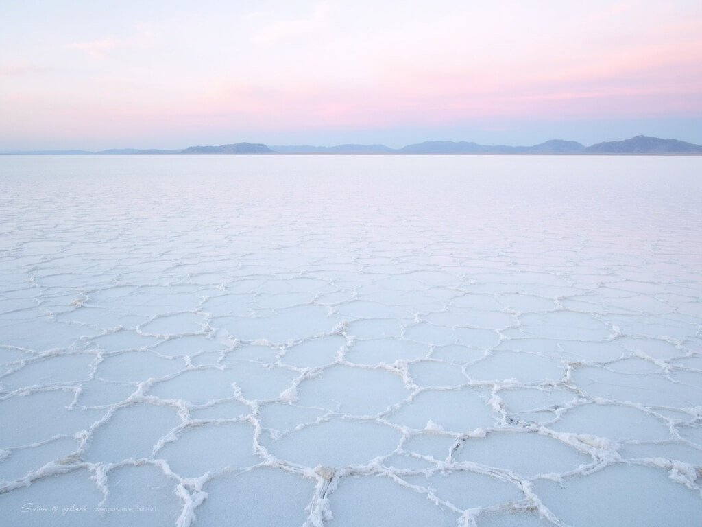 Panoramic dawn view of geometric salt patterns on a flat reflecting the pastel sky with distant mountain silhouettes